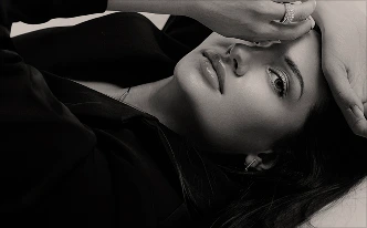 A black-and-white, close-up artistic shot of a woman lying down. Her eyes are open, looking directly at the camera, while her hand—adorned with a shimmering ring—is draped across her forehead.
