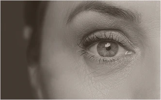 A monochromatic, tight macro shot of a human eye. The focus is on the iris and the fine texture of the skin surrounding the eye, including the eyelashes and subtle expression lines.