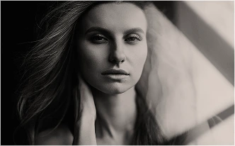 A black-and-white portrait of a woman with long, flowing hair looking directly at the camera with a neutral, soulful expression. She has one hand placed gently against her neck, with soft light hitting the side of her face.