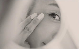 A close-up, high-key black-and-white image of a woman’s eye and cheek. Two fingers with light-colored nail polish are gently touching the skin near the temple, emphasizing skin elasticity or texture.