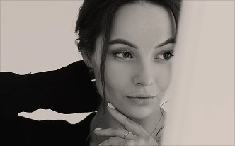 A black-and-white photograph of a woman looking thoughtfully to the side. She is wearing a black long-sleeved top and has one hand tucked behind her head and the other resting on her chin, showcasing her profile and elegant pose.