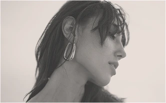 A black-and-white, profile-view photograph of a woman with long, dark hair. She is wearing a large, polished silver hoop earring. Her gaze is directed downward, and the lighting is soft and ethereal against a bright, hazy background.