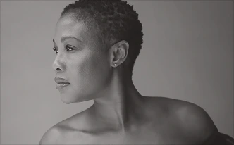 A black-and-white profile portrait of a Black woman with short, textured natural hair. She has a refined profile and is wearing a small stud earring. The lighting highlights the smooth texture of her skin and the definition of her jawline.