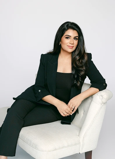 Dr. Sahitya Reddy sitting gracefully in a white tufted armchair. She is dressed in a professional black suit and looking toward the camera with a friendly expression.