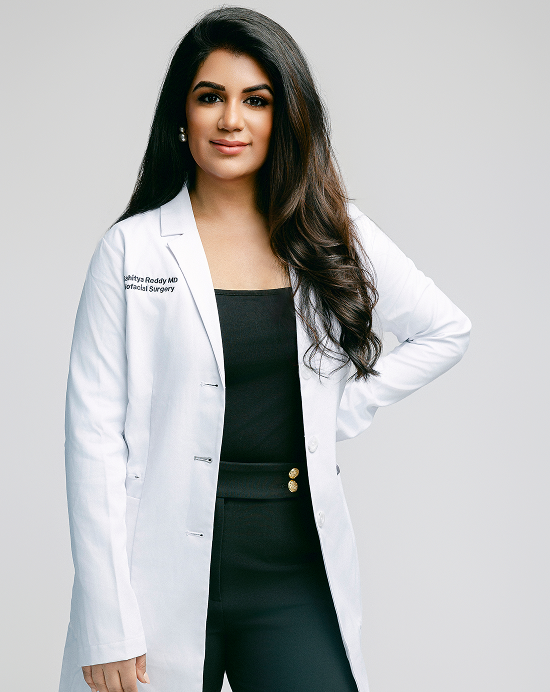 A professional portrait of Dr. Sahitya Reddy, MD, an Oculofacial Surgeon, wearing a white medical lab coat over a black top and trousers. She is standing against a neutral light grey background with one hand on her hip.