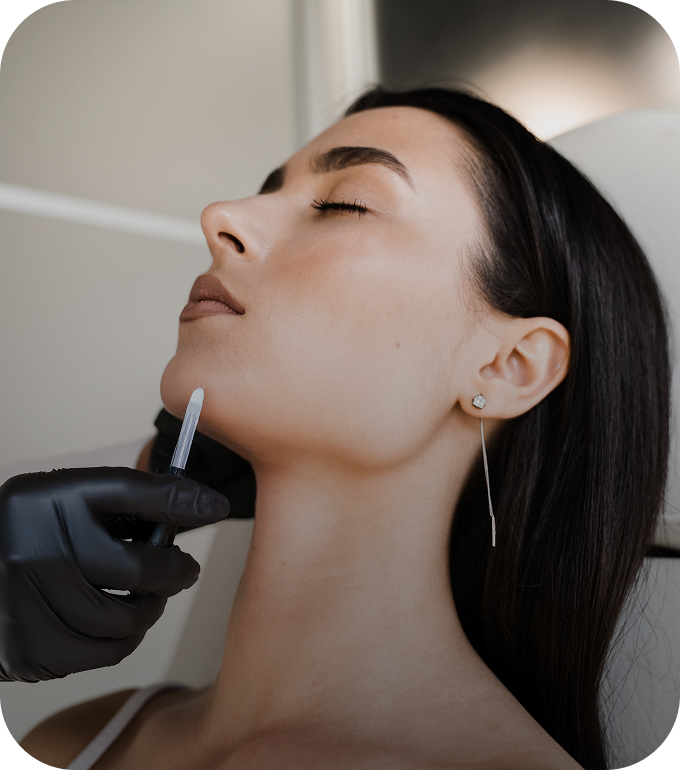 A close-up shot of a woman with her eyes closed receiving a cosmetic injection. A person wearing a black gloved hand is holding a small syringe-like applicator to the underside of her chin.