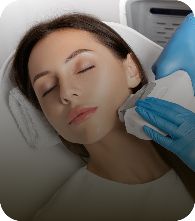 A woman lying down with her eyes closed while undergoing a professional skin treatment. A practitioner in blue gloves is pressing a handheld laser or light-therapy device against her cheek.