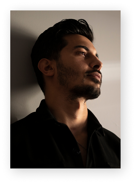 A dramatic, low-light profile portrait of a man with dark, styled hair and a short beard. He is looking slightly upward, with light hitting one side of his face while the rest remains in deep shadow.