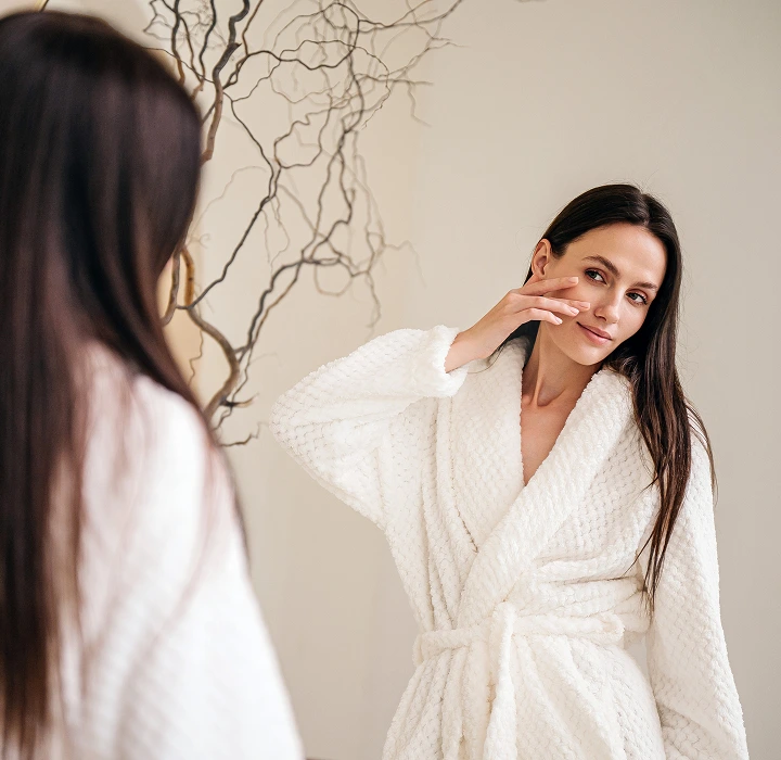 A woman in a white textured bathrobe looking into a mirror and gently touching her face, suggesting a post-treatment or self-care routine.
