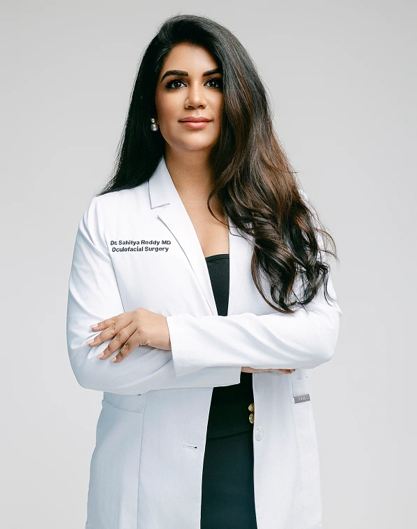 Dr. Sahitya Reddy, MD, Oculofacial Surgeon, posing professionally with her arms crossed. She is wearing a white lab coat with her name and title embroidered on the chest.