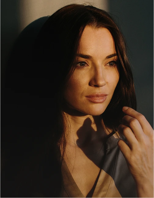 A cinematic portrait of a woman with dark hair, captured in high-contrast "golden hour" light. Half of her face is illuminated by warm sunlight, while the other half is in shadow as she looks thoughtfully to the side.