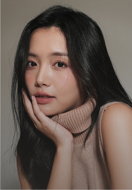 A portrait of an East Asian woman with long, straight black hair wearing a beige turtleneck. She has a gentle expression and is resting her face against her hand, looking directly at the viewer with a soft-focus background.