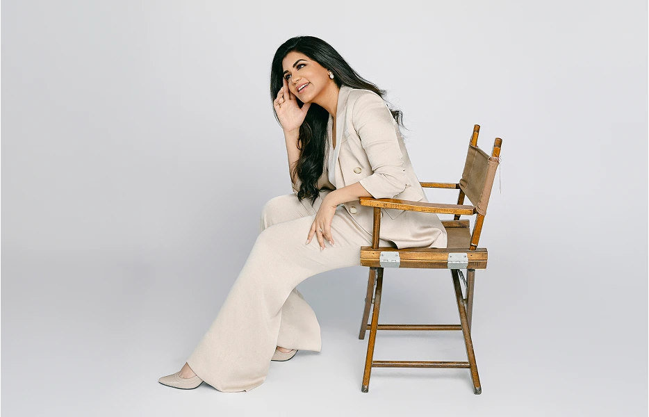 Dr. Reddy sits in a wooden director's chair against a plain grey background, looking off-camera with a bright, engaged expression. She is dressed in a professional beige blazer and matching trousers, resting one hand near her face as if in mid-conversation.