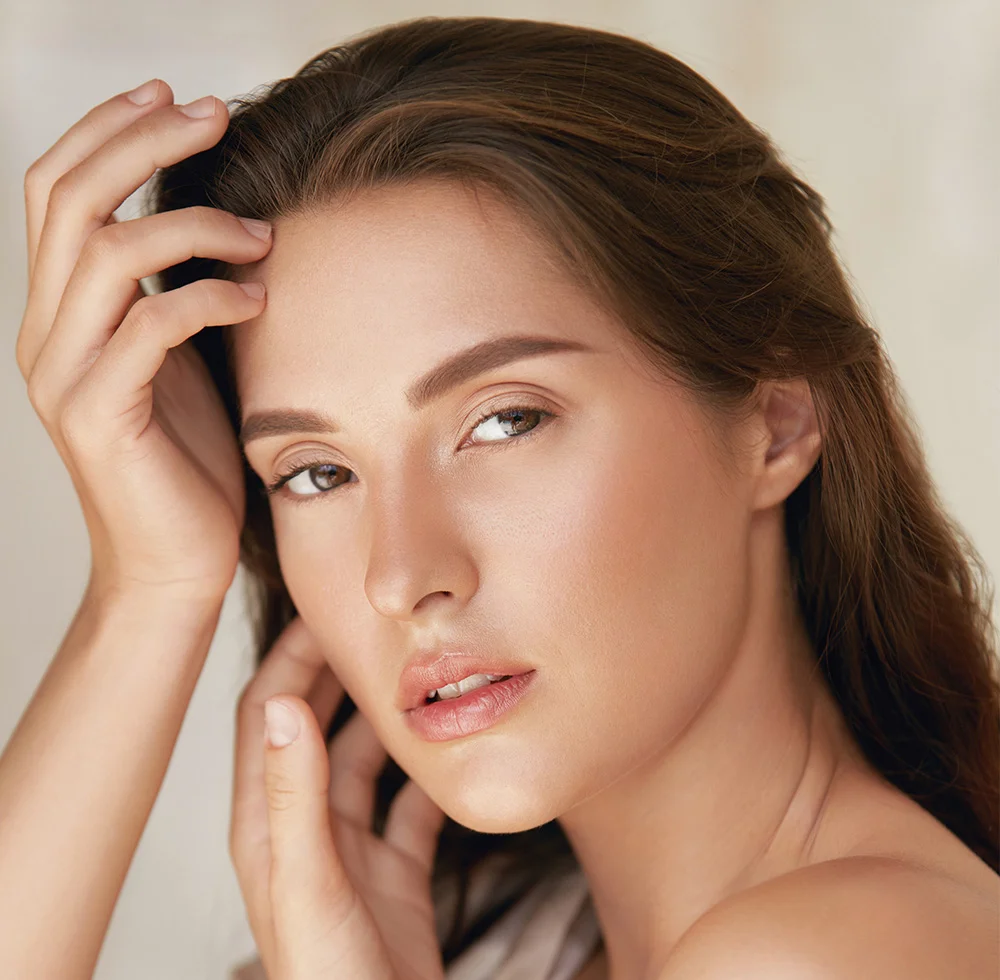 Close up of a young woman with shoulder-length brown hair gazes softly at the camera, her hands gently resting on her head and face. She wears a loose, off-white, off-the-shoulder top. Her smooth, light-toned skin glows under soft, diffused lighting. The muted beige background is softly blurred, keeping the focus on her serene expression and natural beauty. - Almond Eye Lift Canthal Correction Surgery in Draper, UT