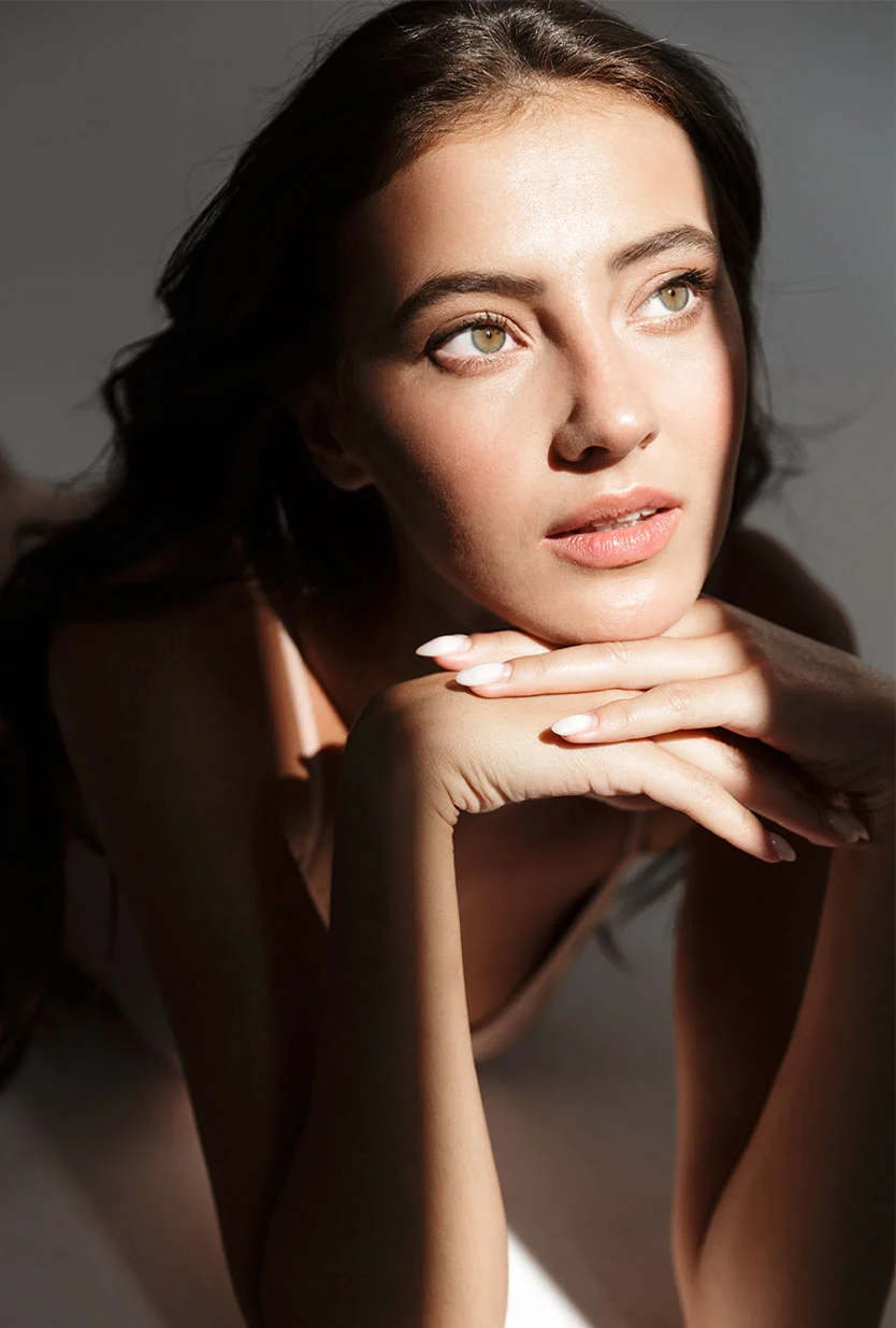 Close-up portrait of a woman with long, dark brown hair lying down, her head resting gently on her hands. She has smooth, light-toned skin, light-colored eyes, and subtle, natural makeup. Her hands are delicately positioned, with nails painted a light, nearly white shade and shaped to a slight point. Soft, dramatic shadows from above create depth and highlight the contours of her face and body. She wears a light pale-beige or nude-colored top. The image emphasizes beauty and the interplay of light on skin. - Almond Eye Lift Canthal Correction Surgery in Draper, UT