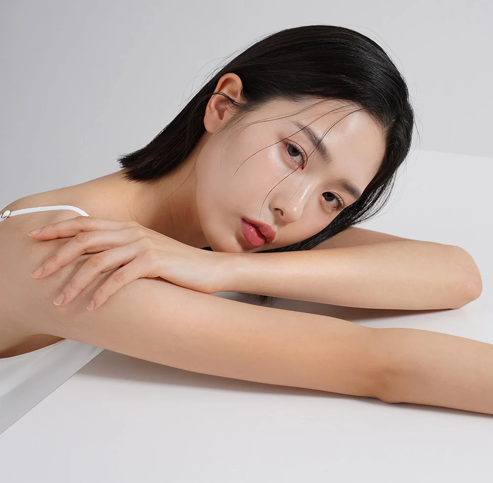 A young Asian woman with a bob haircut, wearing a white spaghetti strap top. She is leaning on a white table with her arms crossed and her head tilted, looking directly at the camera. - Asian Eyelid Surgery in Draper, UT