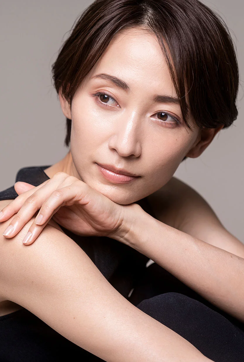A young Asian woman with short, brown hair, wearing a sleeveless black top. She is looking to her left, with her arms crossed and her chin resting on her hands. The background is a solid, light tan. - Asian Eyelid Surgery in Draper, UT