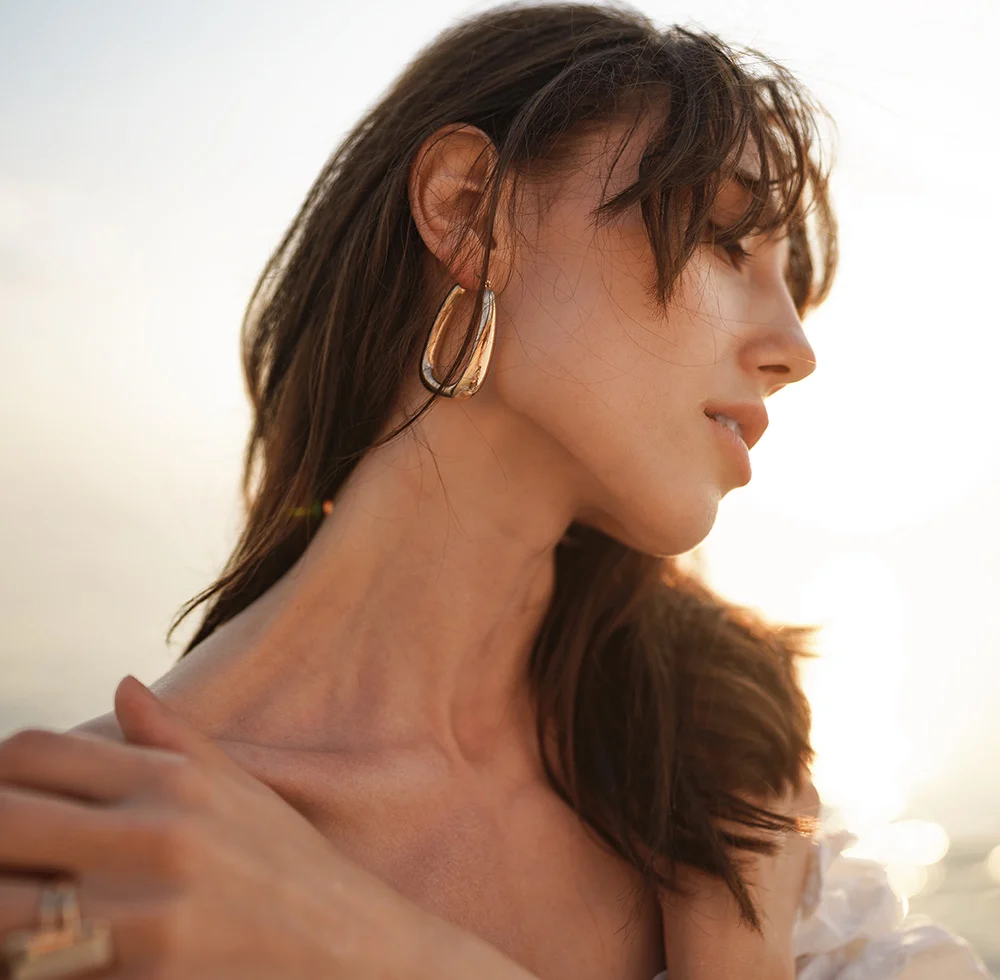 A close up of a woman with dark brown hair from the shoulders and up. She is wearing big gold earrings as she glances down away from camera. - Botox for Neckbands in Draper, UT