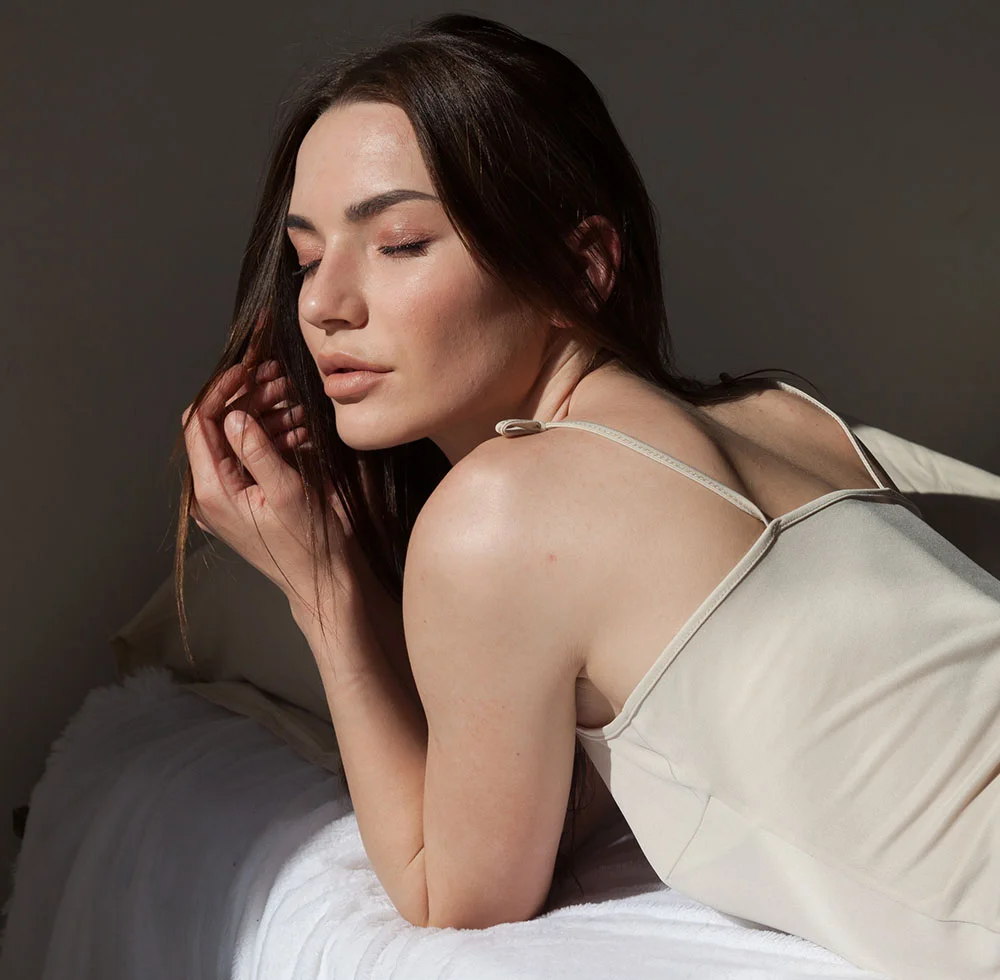 A light-skinned woman with brown hair lies on a bed, her head resting on her hand and her gaze directed off to the side with a pensive or slightly concerned expression. She wears a light beige or cream-colored sleeveless slip dress. Soft lighting casts gentle shadows that highlight the curves of her body, creating an intimate, contemplative mood. White sheets and pillows are visible behind her, with a dim, neutral gray background completing the scene. - Broadband BBL Laser in Draper, UT