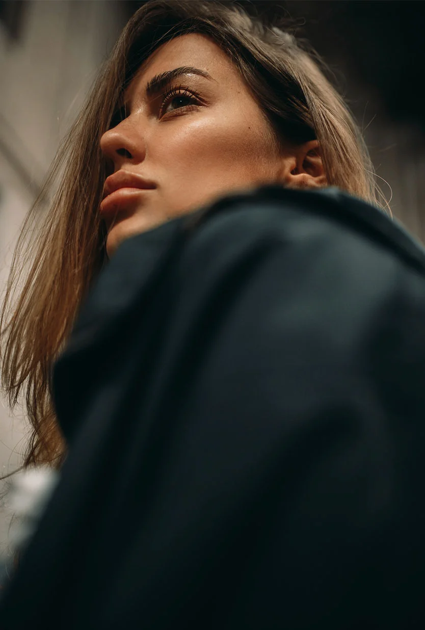 A young woman with a long brown hair, wearing a white button-down shirt. She is seated, looking at her reflection in a mirror, and touching her face with her right hand. The background is a solid, light gray. - Browlift in Draper, UT