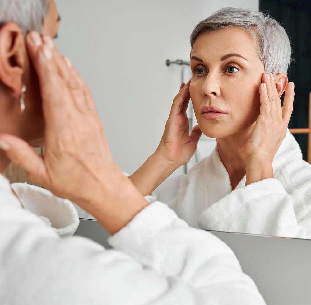 A woman with short, gray hair is looking at her reflection in a mirror. She is wearing a white bathrobe. She is touching her face, and her expression appears somewhat concerned or pensive. The lighting in the bathroom is bright and neutral. The background is a light gray/white bathroom wall. The reflection in the mirror shows a similar-aged woman with a similar expression. - Co2 Laser Resurfacing in Draper, UT