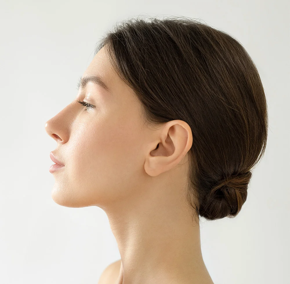 A woman with long, dark hair, wearing a sleeveless white tank top. She is seated on a white bench, looking up and to her right, with her right hand near her lips. The background is a solid light gray. - Chin Augmentation Implant in Draper, UT