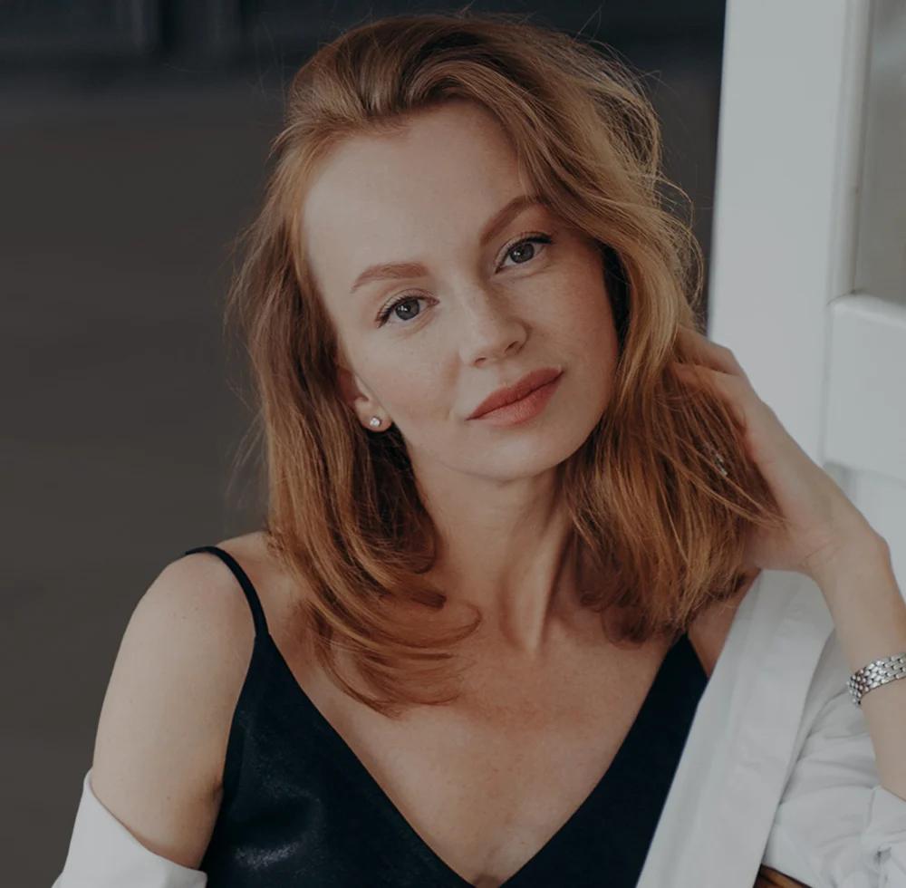 A woman with a short, curly, blonde hairstyle and a bare shoulder, sitting in a dark room with a window, looking up and to her left. She has her hand on her cheek, with sunlight and a shadow on the left side of her face. - Deep Plane Facelift in Draper, UT