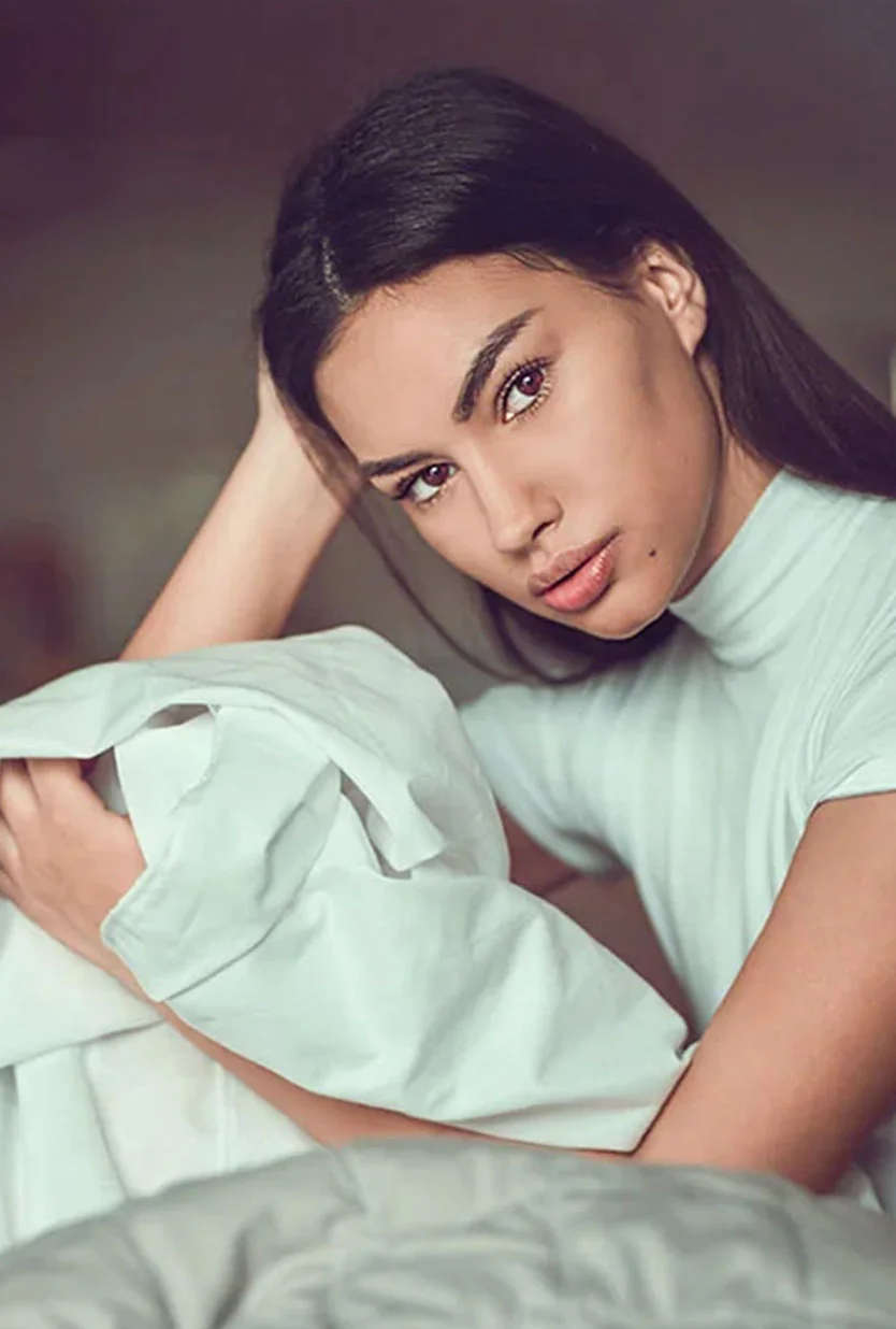 A young woman with long dark hair sits on a bed draped in a white sheet. She looks thoughtfully at the camera, her hands resting on the fabric wrapped loosely around her. She wears a light cream or white turtleneck top. The background is a muted grayish-brown, and soft, diffused lighting highlights her natural complexion in a bedroom setting. - Dermal Fillers in Draper, UT