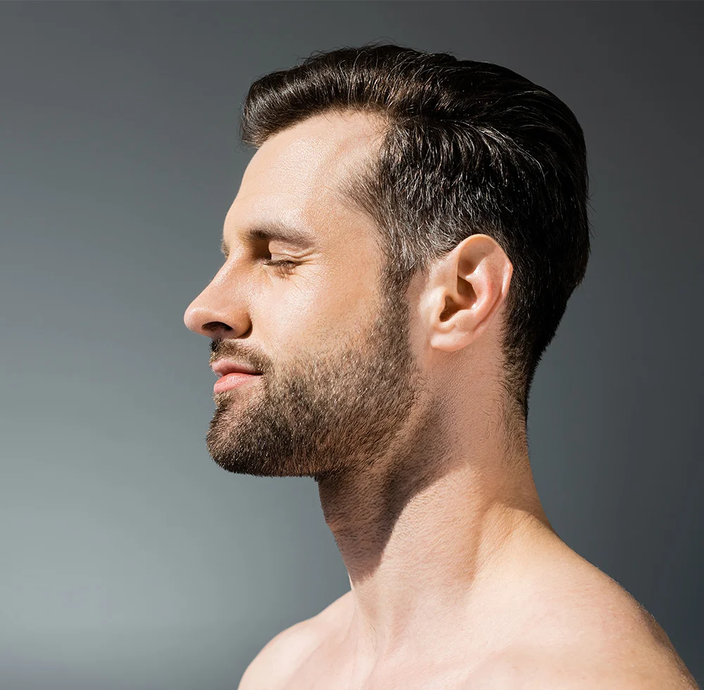 Profile view of a light-skinned, shirtless man with short, neatly styled dark hair and a short trimmed beard. His eyes are closed, and his expression is neutral and contemplative. His chest and shoulders show smooth, light skin with fine chest hair. The muted gray background features shadows cast by a light source, highlighting the contours of his face and body. - Earlobe Repair in Draper, UT