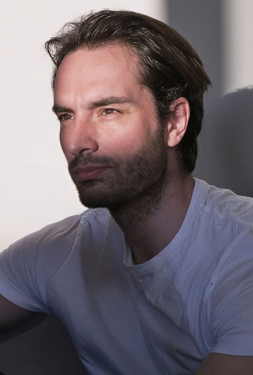 Medium shot of a light-skinned man with short brown hair and a trimmed beard, seated and looking thoughtfully to his right. He wears a plain white t-shirt. The background is a muted grey-blue wall with soft, diffused striped shadows cast by natural light from above left. - Earlobe Repair in Draper, UT