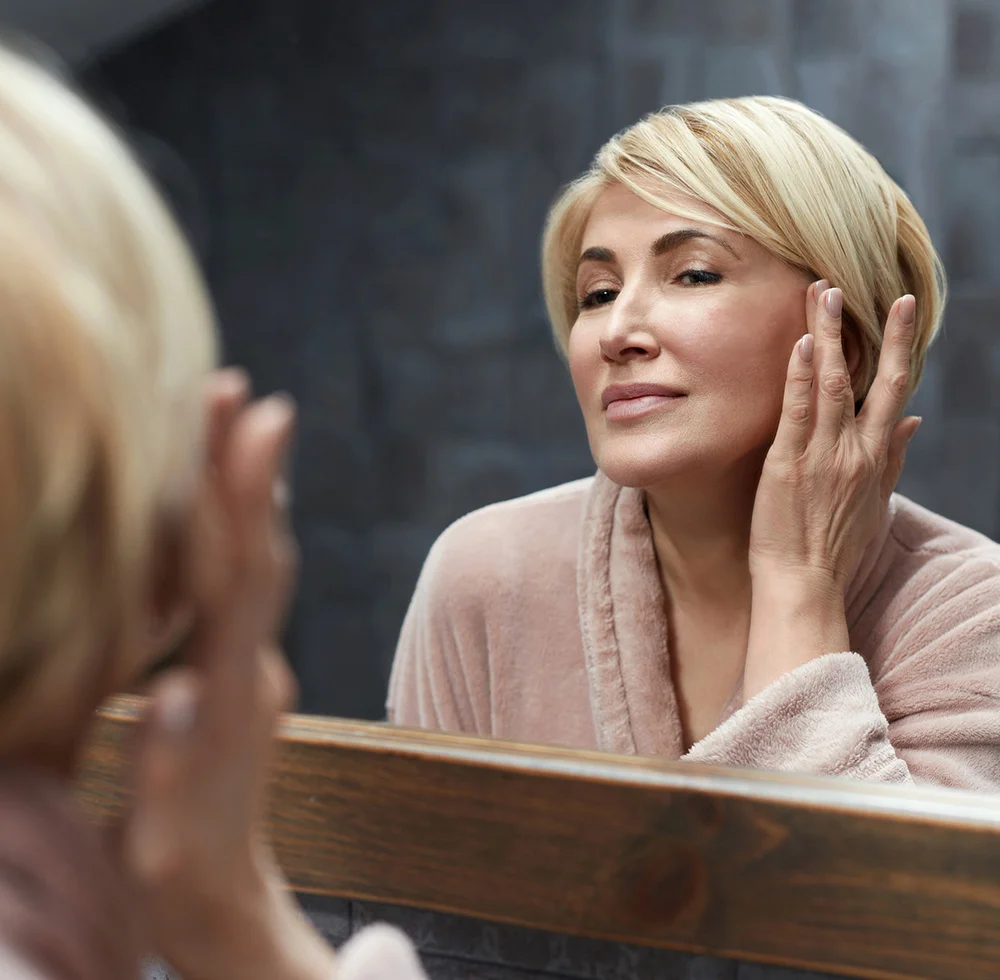 A blonde woman with a short haircut, wearing a pink robe, is looking at her reflection in a mirror. She is touching the left side of her face with her left hand. The mirror has a thick wooden frame, and the background is a dark gray, tiled wall. - Ectropian Repair in Draper, UT