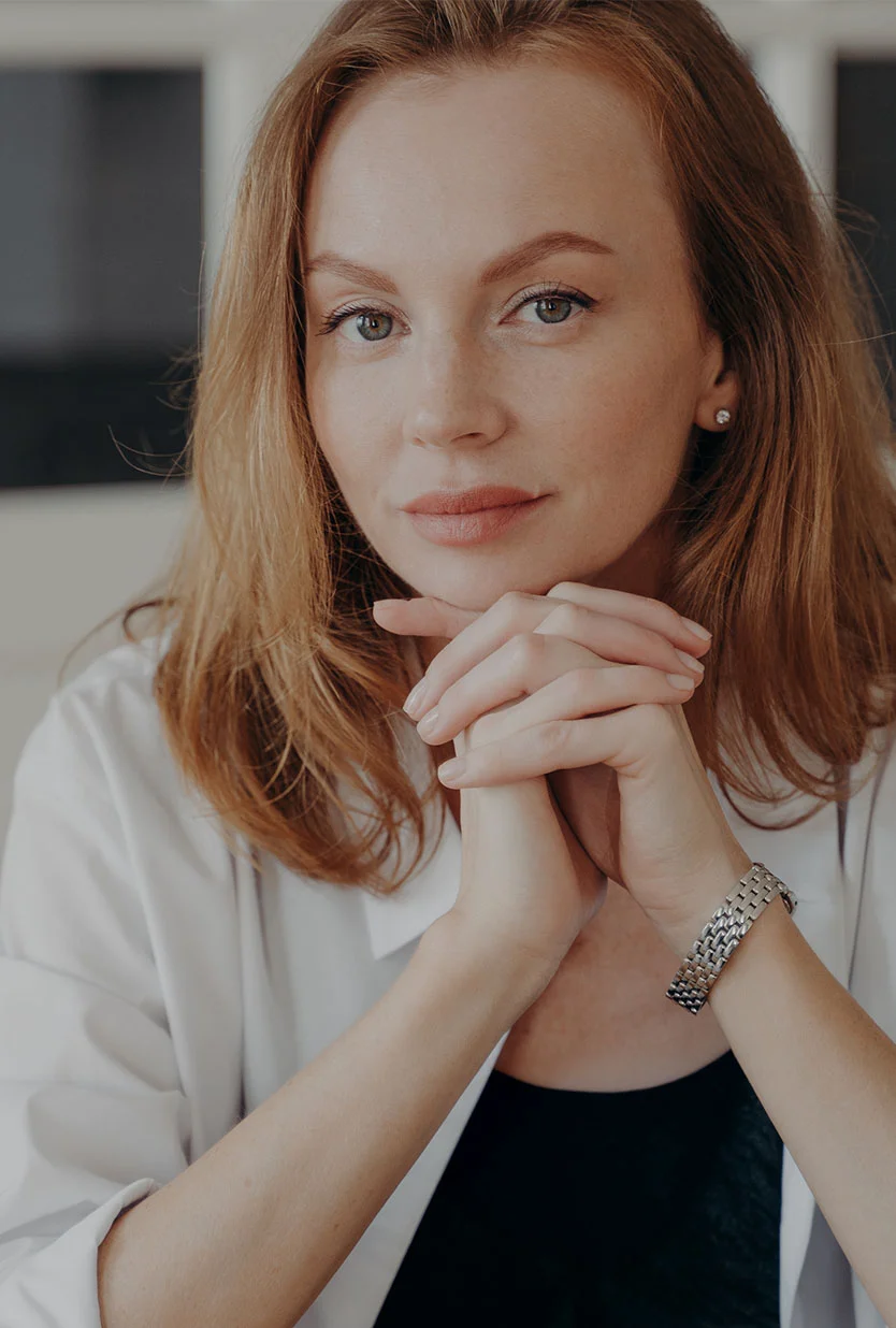 A light-skinned woman with shoulder-length reddish-brown hair is seated, looking directly at the camera with a thoughtful or serious expression. Her light-colored eyes are focused, and her hands are clasped in front of her chin. She wears a white long-sleeved button-up shirt over a dark top, with a silver-toned watch on her right wrist. The softly lit background is out of focus, featuring off-white walls or a light-colored room. - Ectropian Repair in Draper, UT