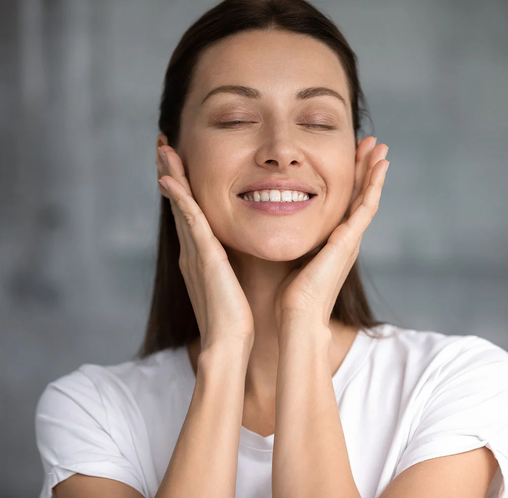 A beautiful woman with her eyes closed, smiling, and touching both cheeks with her hands. She has long brown hair and is wearing a white t-shirt. The background is a solid, light gray. - Erbium Yag Laser in Draper, UT
