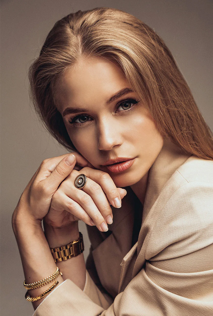 A young woman with long, blonde hair, wearing a beige blazer. She is seated, looking directly at the camera with her hands clasped together near her chin. She is wearing a gold watch, two gold bracelets, and a ring with a large black and gold design. The background is a solid, light gray. - Erbium Yag Laser in Draper, UT
