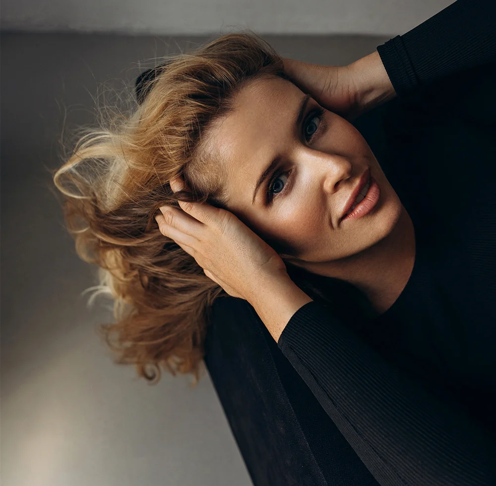 A woman with light brown, tousled hair is viewed from an overhead, slightly angled perspective. She holds her hair with both hands, her head tilted back slightly, and her expression relaxed. She has light skin and light blue eyes. She wears a fitted black top, and the background is a muted gray surface with a soft-focus effect. - Eyelid Retraction Repair in Draper, UT