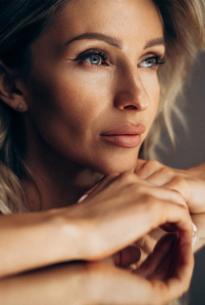 A close-up of a woman with blonde hair and blue eyes, looking to her right. She is resting her chin on her clasped hands and is wearing small stud earrings. The background is a solid, dark gray. - Eyelid Retraction Repair in Draper, UT