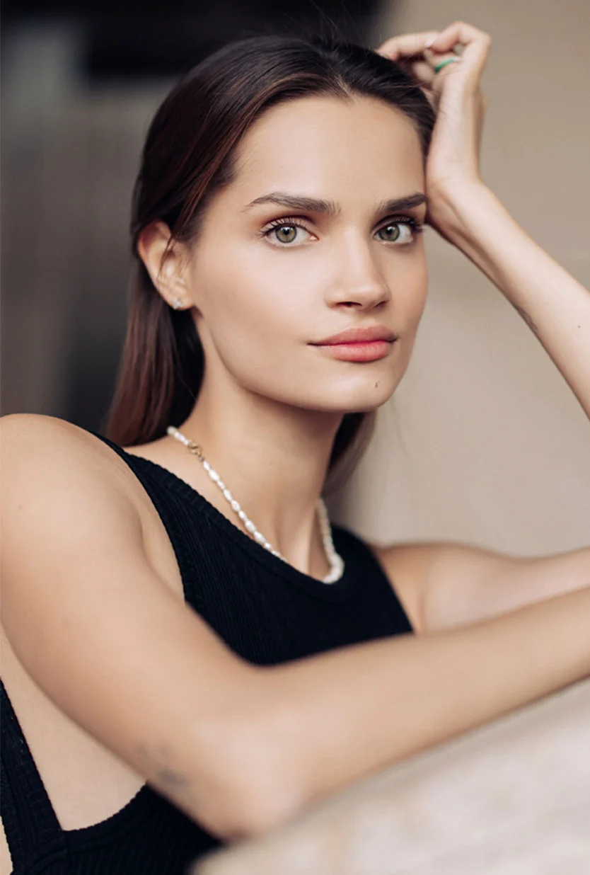 A woman with long, brown hair and light green eyes, wearing a black tank top and a pearl necklace. She is looking directly at the camera with her left arm propped up and her hand resting on her head. The background is a solid, light tan. - Facial Grafting in Draper, UT