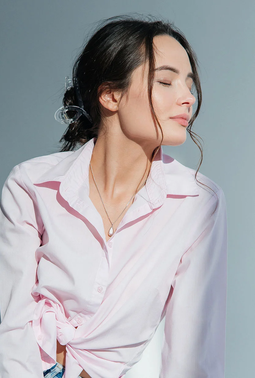 A portrait of a beautiful woman with long brown hair tied up with a clear hair clip, wearing a light pink button-down shirt that is tied at the waist. She has a silver necklace with a teardrop charm and her eyes are closed and her head is tilted up and to the right. The background is a solid, light gray. - Facial Scar Revision in Draper, UT