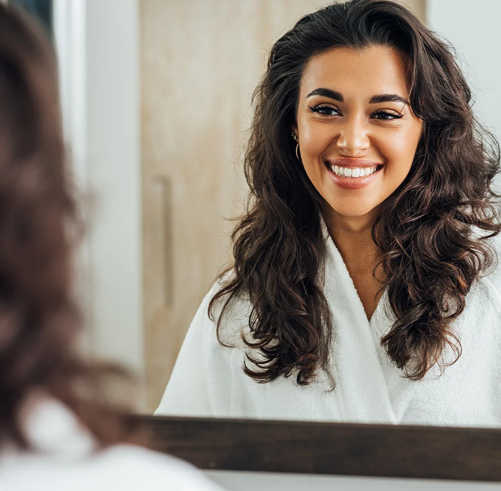 A young woman with long, brown, curly hair, wearing a white robe and smiling at her reflection in a mirror. - Hairline Lowering in Draper, UT