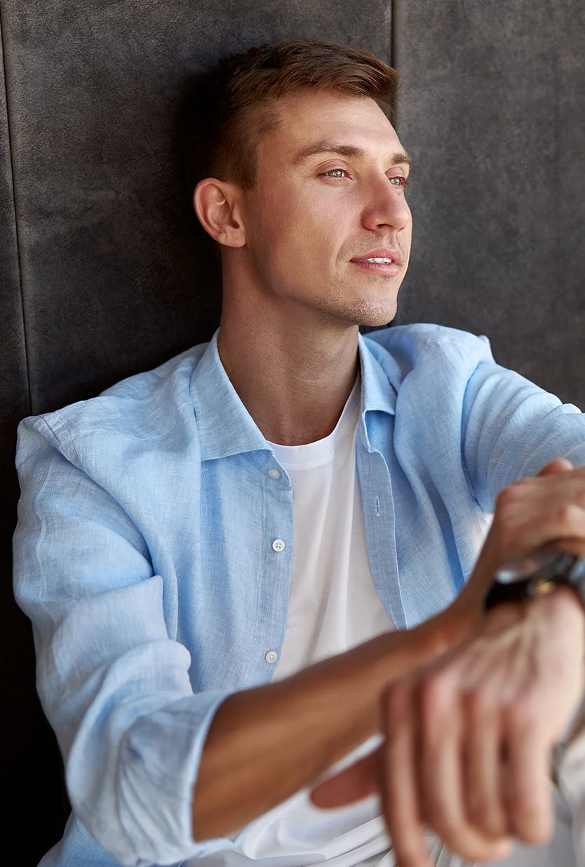 A handsome man with light brown hair, wearing a white t-shirt and a light blue long-sleeved shirt over it, looking to his right. He is seated with his hands clasped, and is wearing a watch with a dark band on his left wrist. The background is a dark gray wall. - Hairline Lowering in Draper, UT