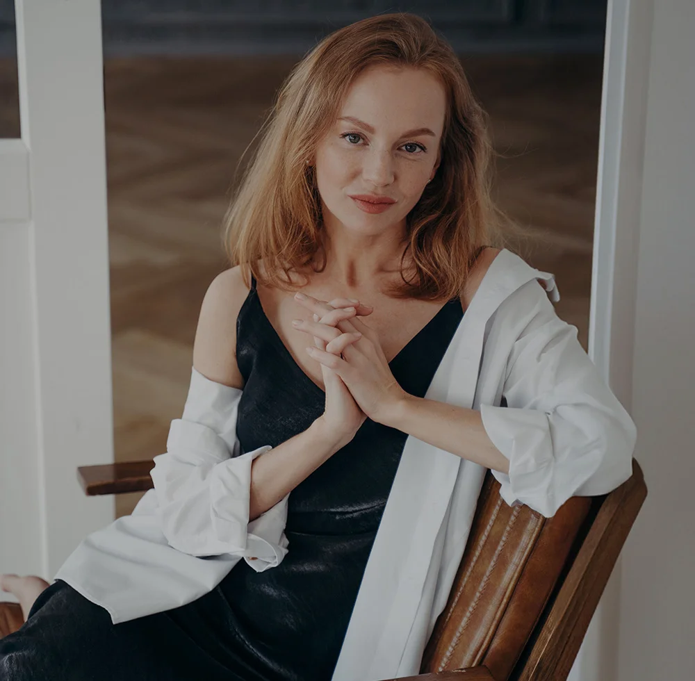 A portrait of a young woman with long, reddish-blonde hair, wearing a black slip dress and a white long-sleeved collared shirt. She is seated in a leather chair, looking directly at the camera with her hands clasped together in her lap. The background is a room with a white door and a hardwood floor. - Hand Rejuvenation in Draper, UT