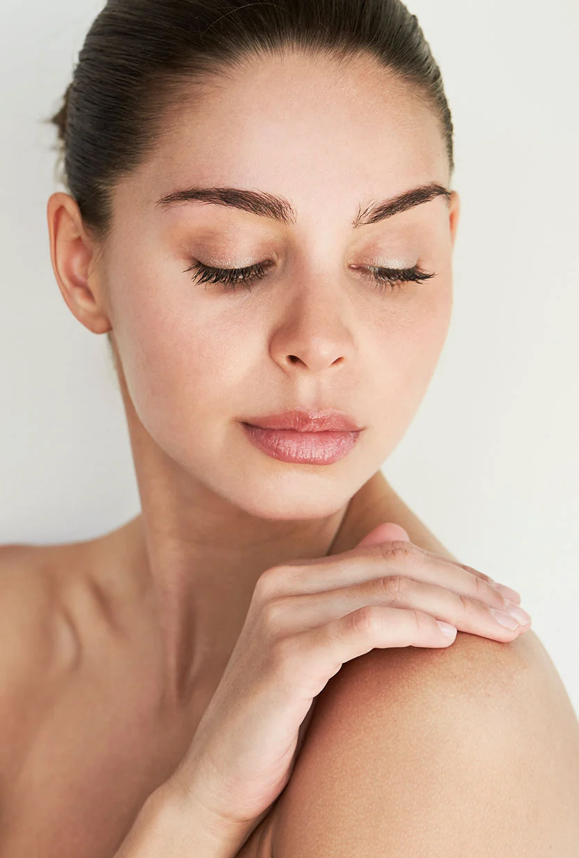 A close-up of a woman with her hair pulled back and her eyes closed, looking down and to her left. She has her left hand resting on her right shoulder. The background is a solid, light color. - Hand Rejuvenation in Draper, UT