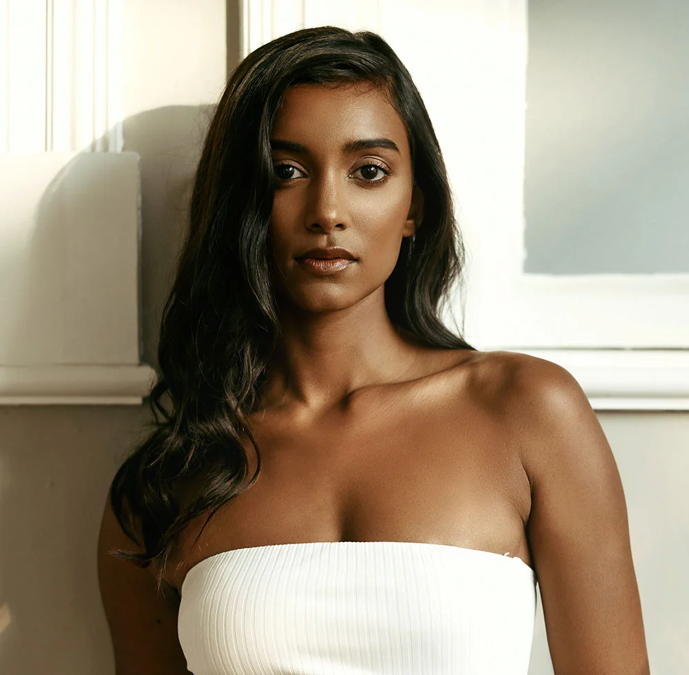 A young woman with long, dark hair, wearing a white strapless top. She is looking directly at the camera with a neutral expression on her face, and sunlight is shining from the left on her face and shoulder. The background is a white wall with a vertical molding. - IPL Intense Pulsed Light Therapy in Draper, UT