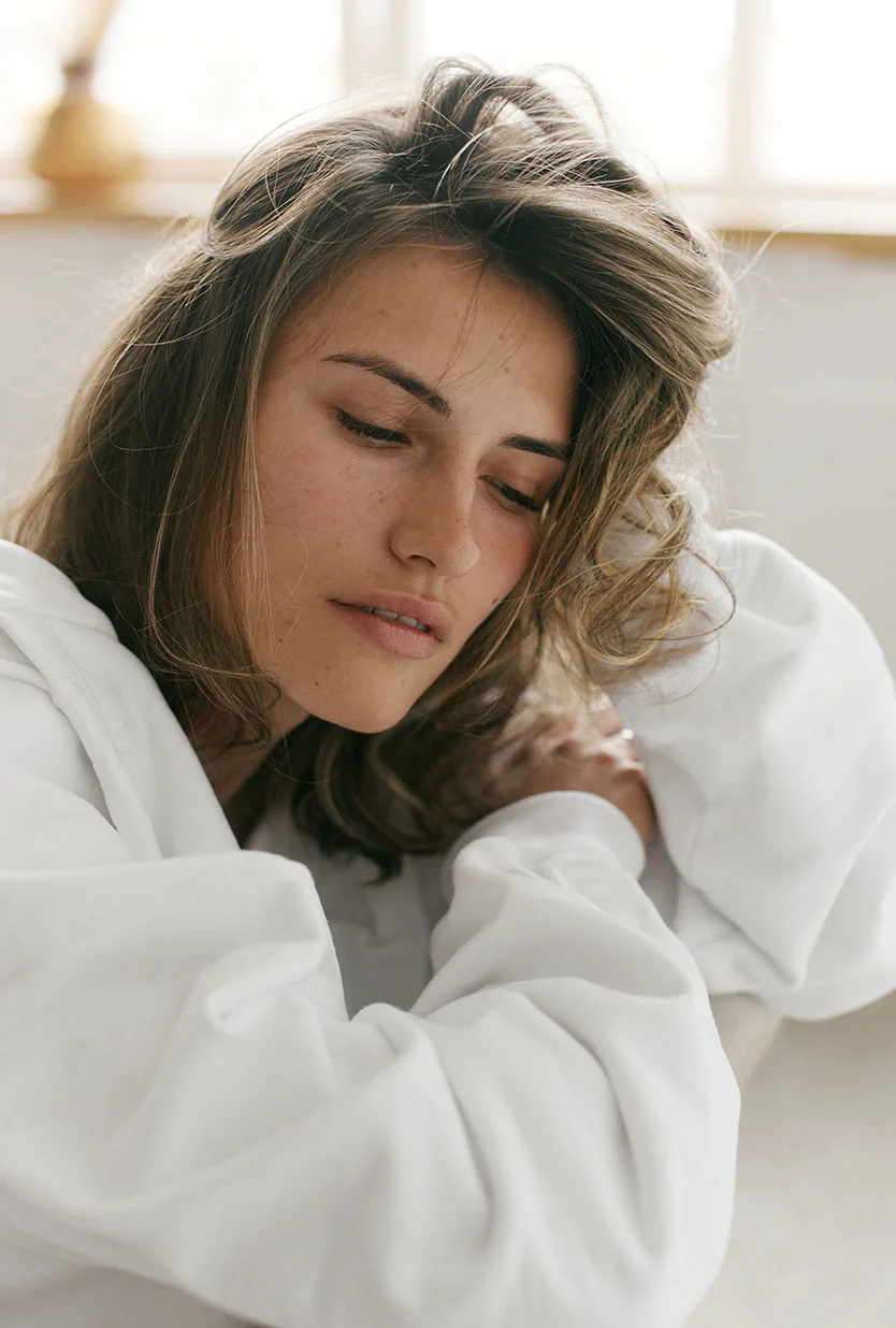 Close-up of a young woman with light brown, slightly tousled hair framing her face. Her expression is neutral and contemplative, with her gaze directed slightly off to the viewer’s right. She has smooth, light-toned skin and wears a white, loose-fitting top. Soft, diffused lighting creates a gentle glow around her, with a window or light source visible in the bright, airy background. - IPL Intense Pulsed Light Therapy in Draper, UT