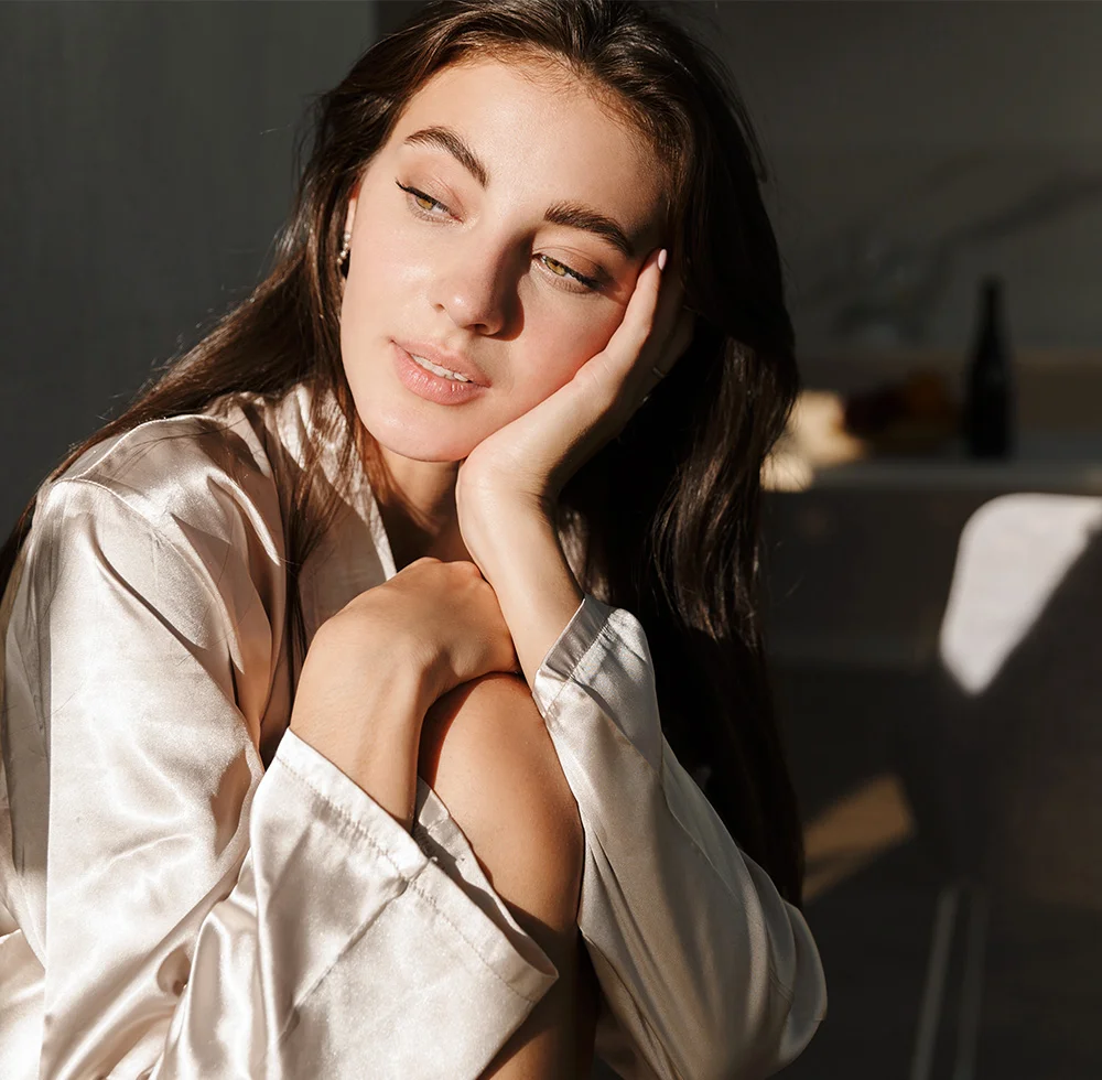 A portrait of a beautiful woman with long brown hair, wearing a light-colored satin robe. She is seated, looking to her left, with her left hand on her cheek and her right arm resting on her knee. Sunlight from the left is shining on her face. The background is a dark, shadowy room. - Jawline Contouring in Draper, UT