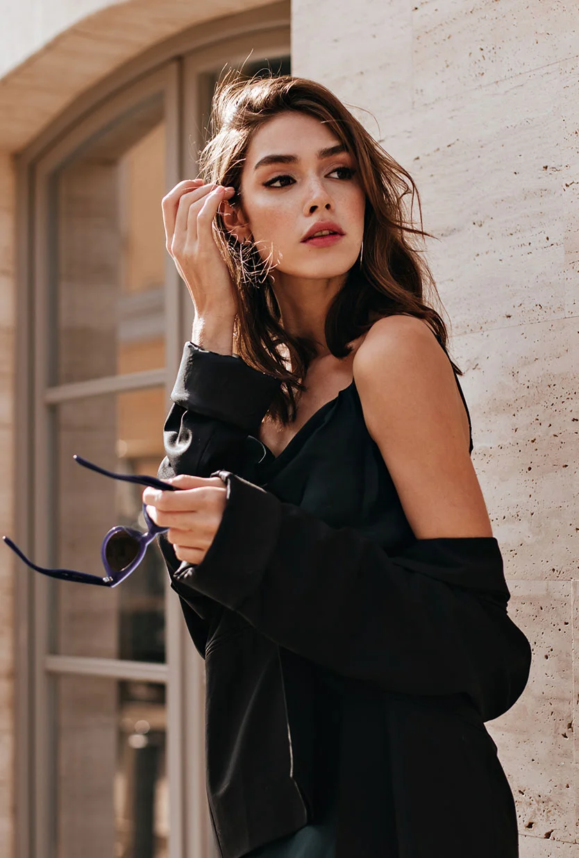 A portrait of a young woman with shoulder-length brown hair, wearing a black slip dress and a black jacket. She is standing next to a stone wall and looking to her right, with her right hand near her temple and holding a pair of sunglasses in her left hand. The background is a window with a rounded arch. - Laser Skin Rejuvenation Deepmediumlight in Draper, UT