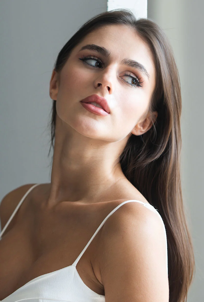 A young woman with long, brown hair, wearing a white spaghetti strap top. She is looking away to her right with a pensive expression, and her hair is draped over her shoulders. The background is a solid gray. - Lip Fillers in Draper, UT