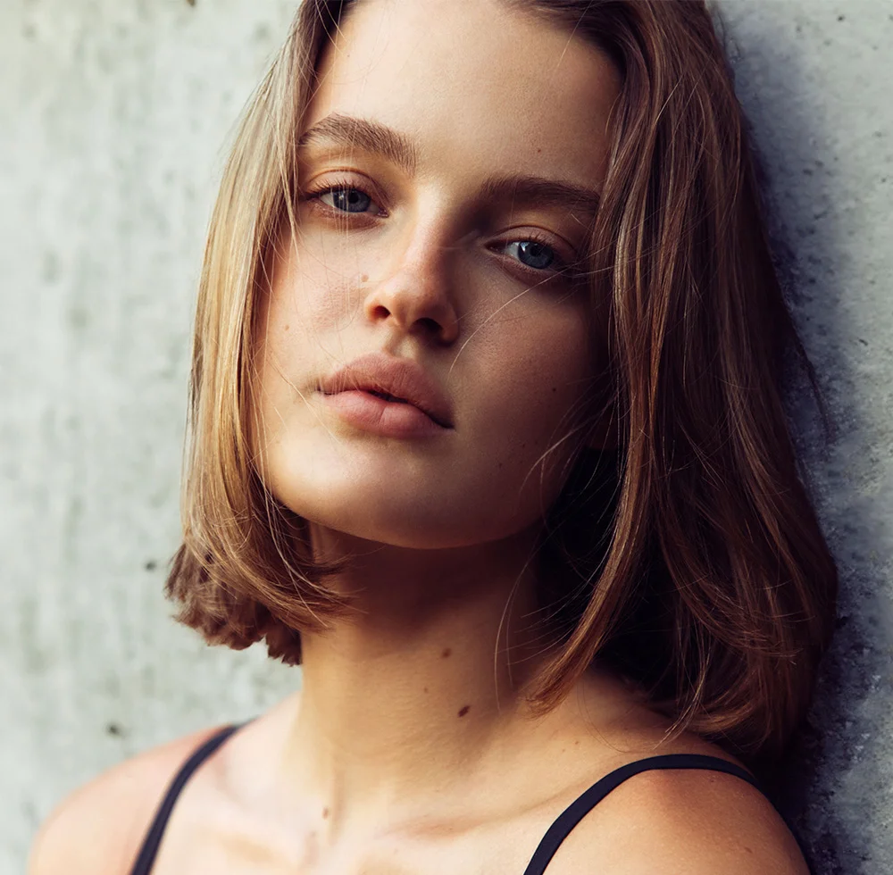 A close-up portrait of a young woman with light brown, shoulder-length hair framing her face. She has smooth, light-toned skin with subtle freckles on her neck and chest. Her neutral expression features soft pink lips and light blue eyes gazing slightly off to the right. She wears a dark sleeveless top against a muted gray-beige background, with natural lighting that highlights her skin’s texture. - Lip Lift in Draper, UT