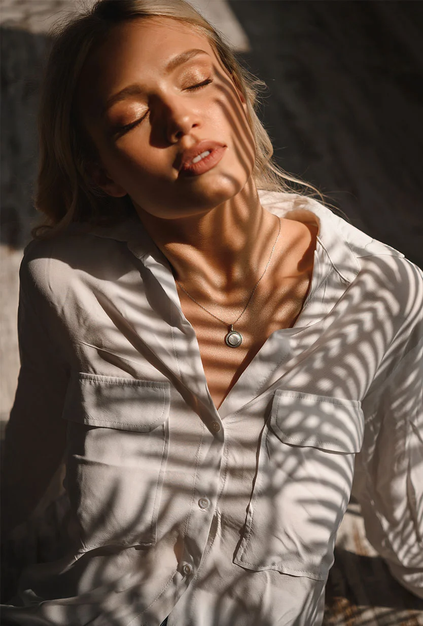 A young woman with light blonde hair reclines with her eyes closed, wearing a loose off-white shirt. Sunlight casts leaf-shaped shadows across her face and body, highlighting her sun-kissed skin. A delicate silver pendant rests at her neck. The muted, textured background and soft lighting create a serene, elegant atmosphere. - Lip Lift in Draper, UT