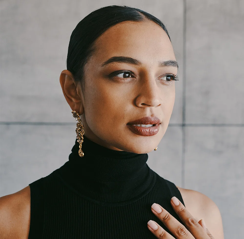 Close-up of a South Asian woman with dark brown skin and shoulder-length hair pulled back. She wears a black turtleneck and gold dangling earrings. Her expression is thoughtful, lips muted reddish-brown, and nails light pink. The background is a neutral light gray stone wall, with soft, even lighting highlighting her features. - Liquid Facelift in Draper, UT